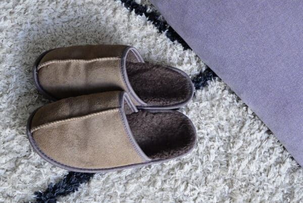 Para beżowych kapci z miękkiego materiału leżąca na puszystym, białym dywanie obok fioletowej poduszki. Osoba zmagająca się z trudnościami, symbolizująca kryzys w procesie zmiany i mentalny opór przed nowym.