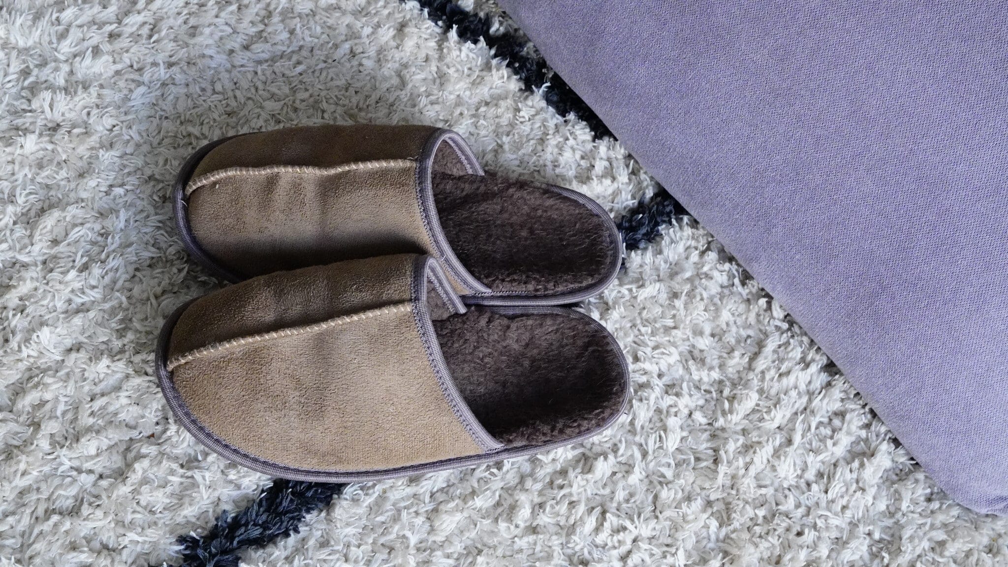 Para beżowych kapci z miękkiego materiału leżąca na puszystym, białym dywanie obok fioletowej poduszki. Osoba zmagająca się z trudnościami, symbolizująca kryzys w procesie zmiany i mentalny opór przed nowym.